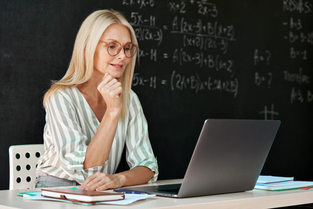 Happy teacher having online webinar maths lesson sitting at chalkboard.