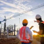 Two engineers from uk construction companies wearing hard hats and safety vests overlooking a construction site with a crane at sunset.
