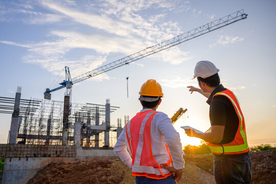 Two engineers from uk construction companies wearing hard hats and safety vests overlooking a construction site with a crane at sunset.