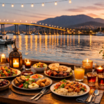 Hobart waterfront at sunset with a table of halal-friendly dishes, lit candles, flowers, and the Tasman Bridge in the background.