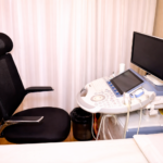 Modern ultrasound examination room with a diagnostic imaging machine, adjustable chair, monitor, and patient bed in a clean clinical setting.