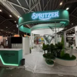 Spritzer exhibition booth with green curved signage, illuminated logo, plants, and seating area inside a convention hall.