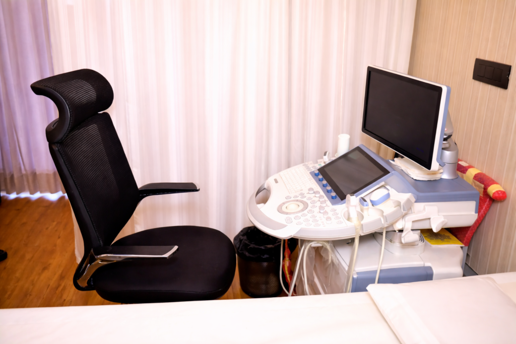 Modern ultrasound machine with monitor and probes beside an adjustable black chair in a clean clinical examination room.