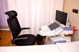 Modern ultrasound machine with monitor and probes beside an adjustable black chair in a clean clinical examination room.
