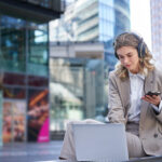 Young businesswoman working on laptop, concentrating in headphones, using her mobile phone, sitting on street of business center