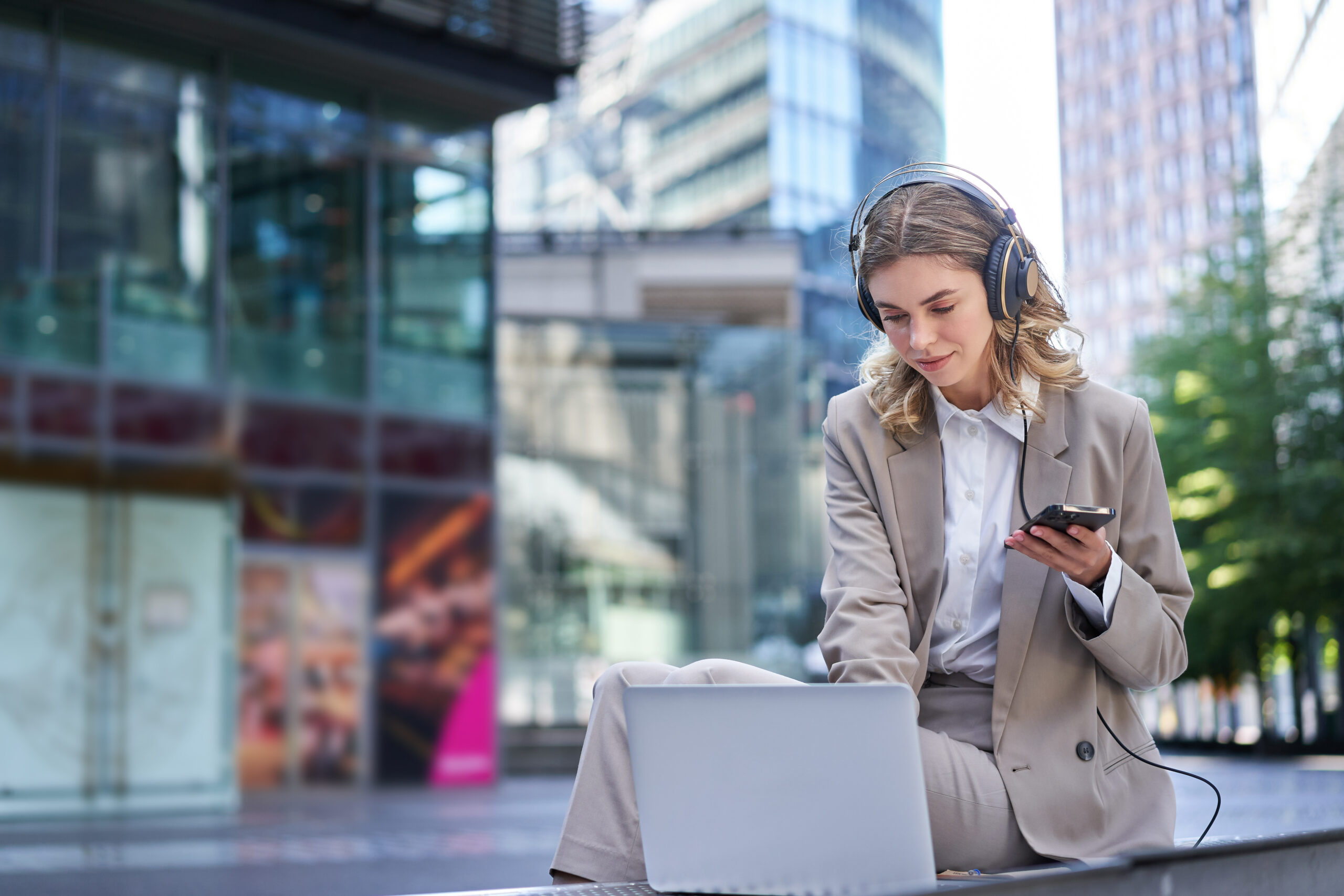 Young businesswoman working on laptop, concentrating in headphones, using her mobile phone, sitting on street of business center